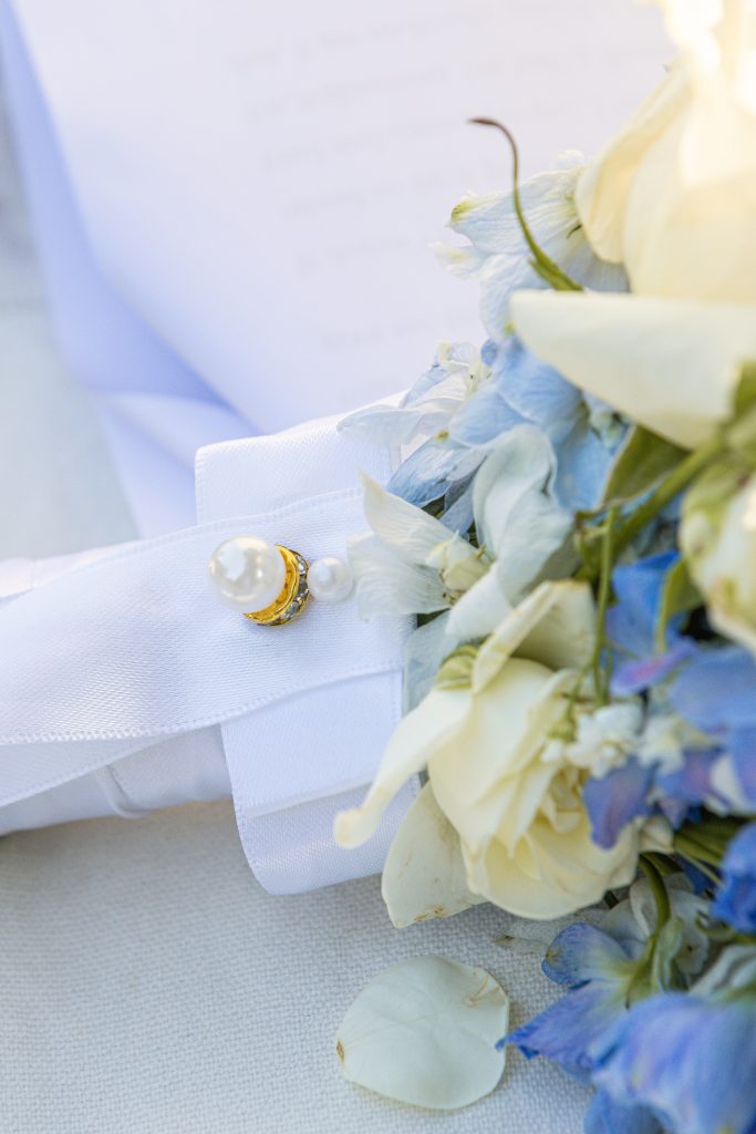 Gros plan sur le ruban d’un bouquet de mariée orné d’une broche perle et dorée, accompagné de fleurs ivoire et bleues.