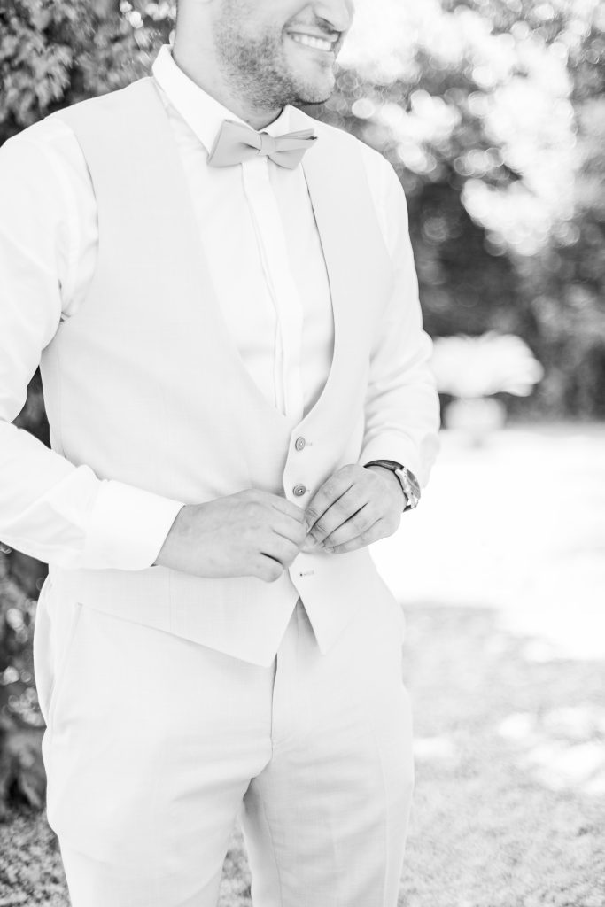 Le marié souriant ajuste son gilet et son nœud papillon, vêtu d’un costume clair en extérieur.