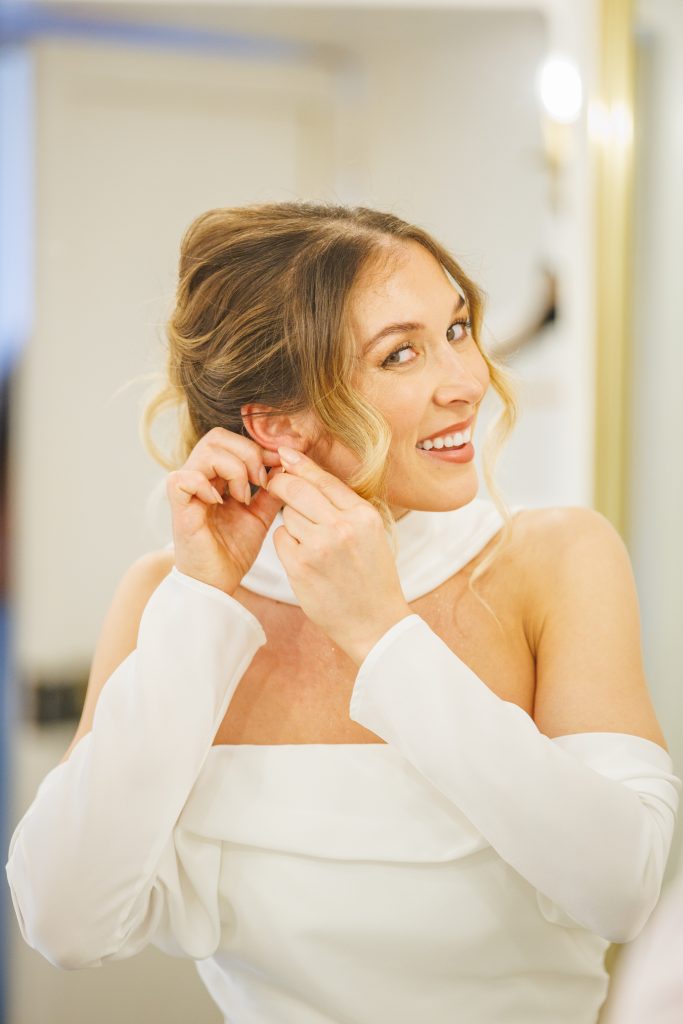 Portrait lumineux d’une mariée souriante ajustant sa boucle d’oreille, dans sa robe blanche élégante.