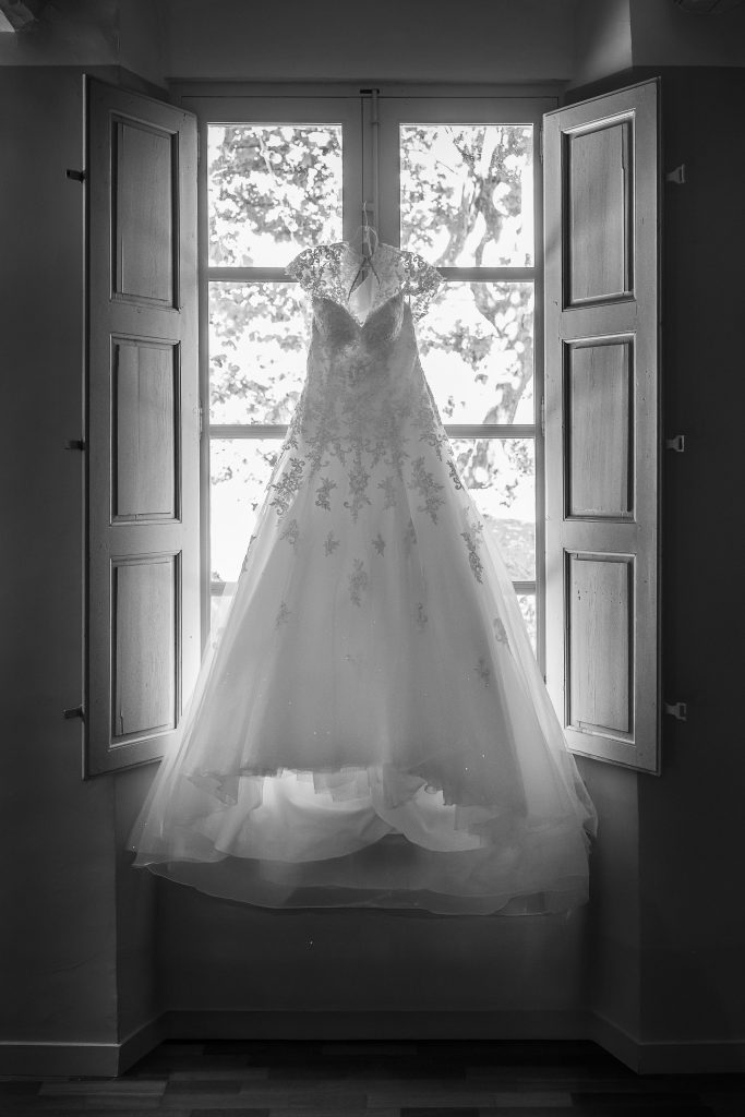 Robe de mariée en dentelle suspendue devant une fenêtre provençale, capturée en noir et blanc par Studio JOLY Photographie, spécialiste du mariage de luxe en Provence.